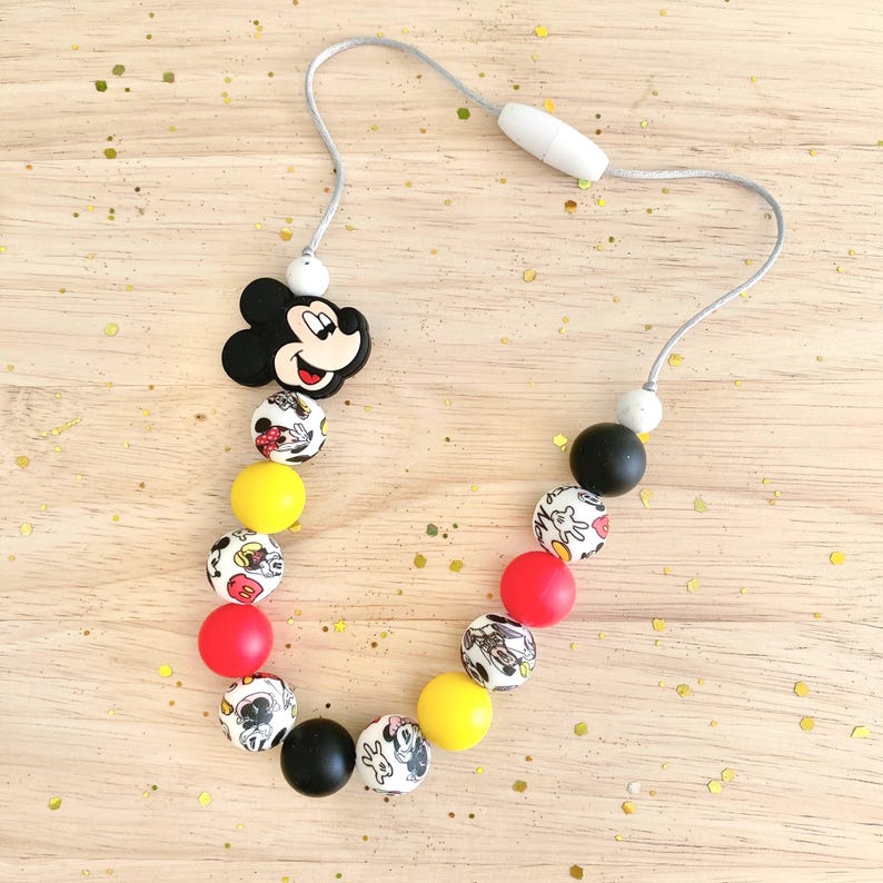 May include: A Mickey Mouse themed necklace featuring a black Mickey Mouse head charm, alternating red, yellow, black, and patterned beads. The necklace is strung on a gray cord with a white clasp.