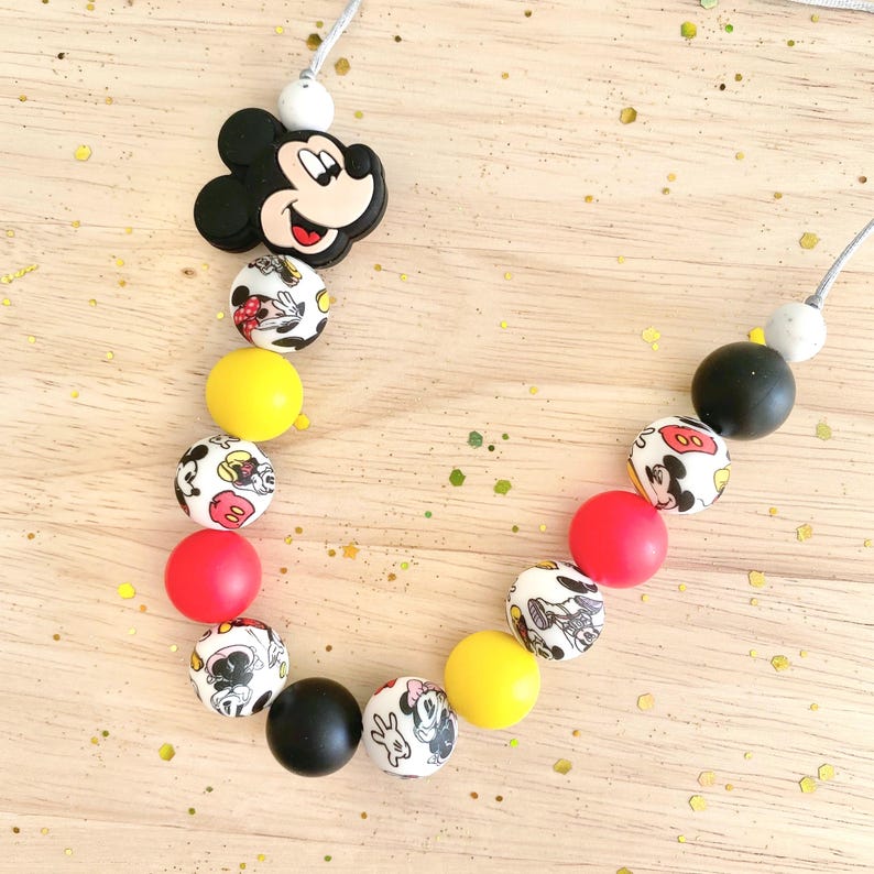 May include: A necklace featuring a black Mickey Mouse head charm and a series of colorful beads. The beads include yellow, red, black, and white with Mickey Mouse designs. The necklace is strung on a gray cord.