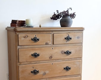 May include: A light wood chest of drawers with five drawers and black metal handles. The chest of drawers has turned legs and sits on a light wood floor.