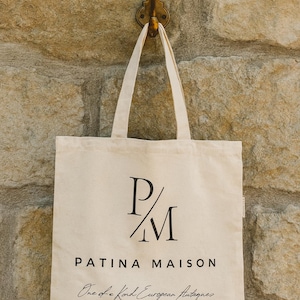 Puede incluir: Bolso tote color crema con texto y asas negras, colgado de un gancho de latón en una pared de piedra. El bolso muestra "P/M PATINA MAISON One of a Kind European Antiques PARKER. CO."
