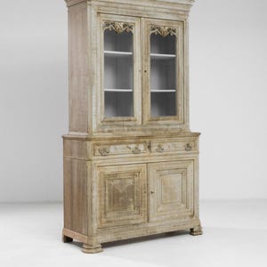 May include: A tall, antique, whitewashed wooden cabinet with two glass doors on top and two solid wood doors on the bottom. The cabinet has two drawers below the top doors and decorative carvings on the top and bottom.