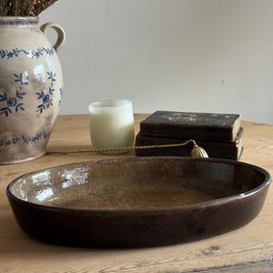 May include: An oval-shaped, dark brown ceramic dish with a glossy, mottled finish. The dish has a shallow, wide basin and smooth, rounded edges, presenting a simple, functional form for holding various items on a flat surface.
