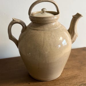 May include: A beige ceramic teapot with a lid and a curved handle. The teapot has a spout and a handle on the side. The surface has a crackled glaze. The teapot is sitting on a wooden surface.