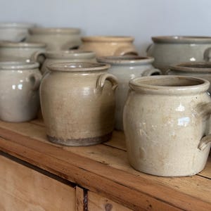 May include: A collection of vintage ceramic crocks in various sizes and shades of beige, displayed on a wooden shelf.