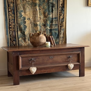 Puede incluir: Un cofre de madera antiguo de color marrón oscuro con dos cajones, adornado con herrajes metálicos y adornos en forma de corazón. El cofre está coronado con un jarrón decorativo, libros y una vela. Una tapicería cuelga en el fondo.