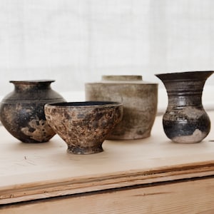 May include: Three ceramic pots in various shades of brown and black, sitting on a light wood surface. The pots are all different shapes and sizes, with one being a wide bowl, one being a tall vase, and one being a shorter, round pot.