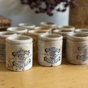 May include: A collection of vintage J. Sainsbury's Potted Meats jars. The jars are cylindrical, with a cream-colored, crackled glaze. Each jar features a black and white label with the brand name and product description. The jars are arranged on a wooden surface.