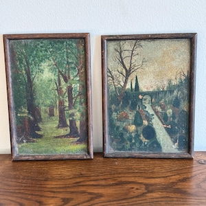 Puede incluir: Dos pinturas de paisajes enmarcadas. Una representa una escena de bosque con árboles altos y follaje verde. La otra muestra un camino que conduce a un puente con árboles y una paleta de colores apagados. Ambas están en marcos de madera oscura.