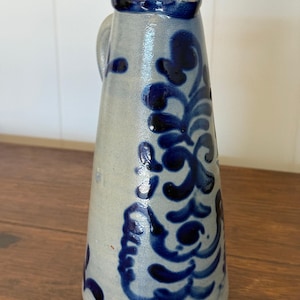 May include: A tall, conical stoneware pitcher with a light gray, salt-glazed finish. The exterior is decorated with hand-painted, deep blue floral and vine motifs. It features a small handle on the side and a simple, unadorned rim.