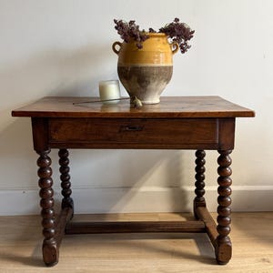 May include: A wooden side table with a square top and turned legs. The table has a drawer and is topped with a decorative vase with dried flowers. A candle and a decorative object are also on the table.