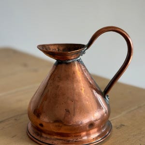 May include: A copper pitcher with a wide, conical body, a flared rim, and a single curved handle attached to the side. The metal surface displays a warm, metallic orange-brown tone with subtle variations in texture and a polished finish.
