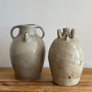 May include: Two antique stoneware jugs with a light beige, speckled glaze. The left jug features two side handles and a small spout. The right jug has a central neck, two small handles, and a small spout. Both vessels have a rounded body shape.