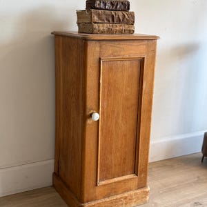 May include: A tall, rectangular wooden cabinet with a light brown finish, featuring a single door with a small, round white knob. Two weathered, leather-bound books are stacked horizontally on the flat top surface of the cabinet.