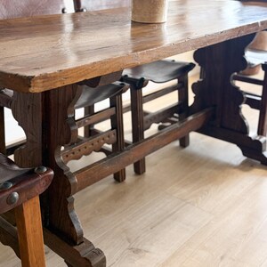 May include: A rustic wooden dining table with a distressed finish, paired with leather-seated chairs. The table features a thick, rectangular top and ornate, carved legs. A small, beige ceramic pot with dried flowers sits on the table.