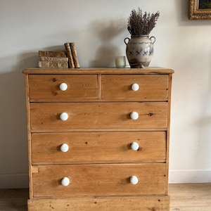 May include: A light brown wooden chest of drawers featuring five drawers arranged in two smaller top drawers and three full-width lower drawers. Each drawer is fitted with a round white knob. The wood grain is visible throughout the structure.