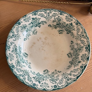 May include: A round ceramic bowl featuring a cream-colored base with a dark green floral transferware pattern. The rim is decorated with an ornate scrollwork border, while the center shows signs of age with light brown discoloration spots.