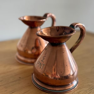 May include: Two copper measuring jugs with a wide flared rim and a curved handle. The front of the foreground jug is engraved with the text 1/2 PINT. Both vessels feature a conical body shape and a polished metallic finish with visible patina.