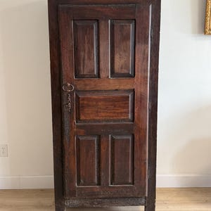 Puede incluir: Un armario alto de madera marrón oscuro con una sola puerta. La puerta está dividida en cuatro paneles rectangulares. El armario tiene una parte superior decorativa y una cerradura de metal. La madera tiene una veta visible y un aspecto envejecido.