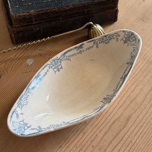 May include: An oval-shaped ceramic gravy boat featuring a cream-colored base with a blue floral and scrollwork pattern along the rim. The piece has a pointed spout and a smooth, glazed finish with visible signs of wear along the edges.