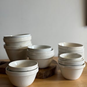 May include: A collection of off-white ceramic bowls of varying sizes, stacked and arranged on a wooden surface. The bowls have a simple, classic design with a slightly flared rim. The neutral color palette and clean lines suggest a minimalist aesthetic.