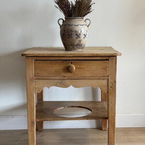 May include: A light brown wooden nightstand with a single drawer featuring a round knob, a scalloped apron, and a lower shelf with a circular cutout. A beige ceramic vase with blue floral patterns and two handles sits on top, filled with dried plants.