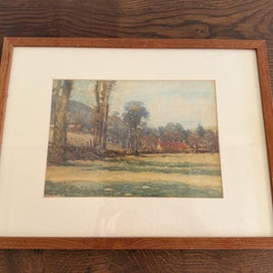Puede incluir: Una pintura de acuarela enmarcada de un paisaje rural. La obra de arte representa un campo, árboles y edificios bajo un cielo azul. La pintura está enmarcada en un passe-partout crema y un marco de madera marrón.