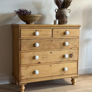 May include: A light brown wooden chest of drawers featuring a natural wood grain finish. The piece has two small top drawers and three full-width lower drawers, all equipped with round white ceramic knobs and small keyhole escutcheons.