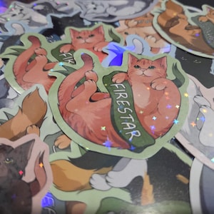May include: A collection of colorful cat stickers. The stickers feature various cat illustrations, including a ginger cat labeled "Firestar." Some stickers have holographic accents. The overall theme is cat-themed merchandise.