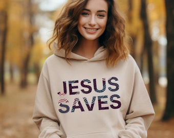 Puede incluir: Una sudadera con capucha beige con el texto "JESUS SAVES" impreso en negro con un patrón colorido superpuesto. El texto está en negrita, sin serif.