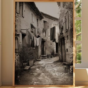 May include: A framed black and white photograph of a narrow European street scene. The image shows stone buildings, a cobblestone street, and laundry hanging from the buildings. The photograph is displayed in a room with a window and plants.