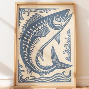 Pode incluir: Uma impressão de arte emoldurada apresentando uma ilustração em linoleogravura azul-escura de um peixe saltando através de ondas estilizadas sobre um fundo creme. A obra está inserida numa moldura de madeira clara de bordas simples.