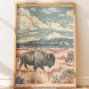 May include: A framed art print featuring a woodcut-style illustration of a dark bison walking through a grassy field. In the background, a snow-capped mountain rises under a blue sky filled with stylized clouds. The color palette includes muted blues, creams, and earthy reddish-brown tones.