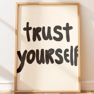 May include: A framed art print with the words "trust yourself" in bold, black, handwritten-style lettering. The background is a light beige, and the frame is a light wood color. The artwork is displayed vertically.