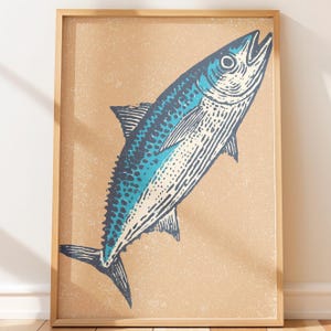 Pode incluir: Uma impressão de arte emoldurada apresentando uma ilustração gráfica estilizada de um peixe. O peixe é renderizado em tons de azul e branco com linhas detalhadas, sobre um fundo bege texturizado dentro de uma moldura de madeira clara.