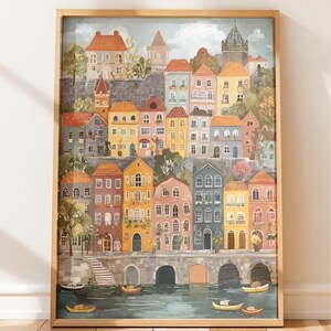 Könnte beinhalten: Ein gerahmter Aquarelldruck eines europäischen Dorfes. Das Kunstwerk zeigt farbenfrohe Gebäude an einem Ufer, mit Booten im Wasser und einer Brücke. Die Gebäude sind in Orange-, Gelb-, Rosa- und Blautönen gehalten.