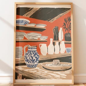 May include: A framed art print featuring a stylized illustration of a kitchen interior with shelves holding various ceramic bowls, pitchers, and vases. The color palette includes shades of terracotta orange, cream, black, and blue with a patterned pitcher.