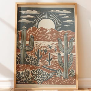 May include: A framed print featuring a desert landscape with cacti, mountains, and a sunburst design. The color palette includes shades of brown, blue, and cream, creating a vintage aesthetic. The artwork is a linocut style print.