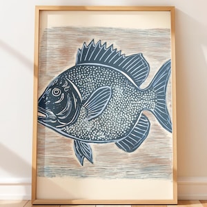 May include: Framed art print featuring a stylized fish illustration in shades of blue and white. The fish is depicted with detailed scales and fins, set against a textured, light brown background. The artwork is encased in a natural wood frame.