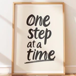 May include: A framed print with the words "One step at a time" in black brush lettering on an off-white background. The frame is made of light brown wood. The artwork is displayed vertically.