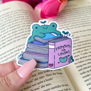 May include: A sticker featuring a teal frog sitting atop a stack of books. The book on top has the title "Froggies to Lovers" and a heart. The sticker is on a book with pink flowers in the background.