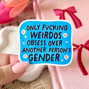 May include: A blue sticker with white text that reads "ONLY FUCKING WEIRDOS OBSESS OVER ANOTHER PERSON'S GENDER" is held in front of a pink book and a bouquet of pink and orange tulips.