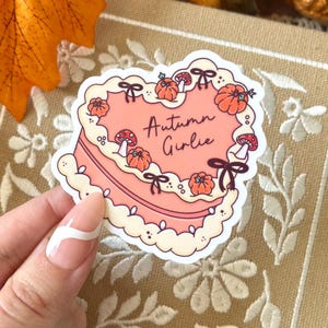 Peut inclure: Un autocollant en forme de gâteau en cœur avec une base de glaçage rose clair et des bords festonnés couleur crème. Le gâteau est décoré de petites citrouilles orange, de champignons rouges et de nœuds marron. Le texte Autumn Girlie est écrit en brun.