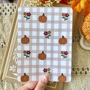 May include: A rectangular card featuring a repeating pattern of orange pumpkins and small clusters of red and brown flowers set against a light beige and white gingham background. The card has rounded corners and a smooth, matte finish.