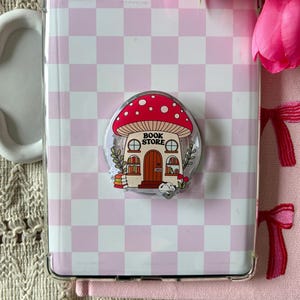 May include: A clear phone case with a light pink and white chequered pattern, featuring a central circular grip attachment decorated with a red mushroom-shaped building labelled BOOK STORE, complete with windows, a door, and small stacks of books.