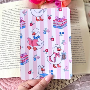 May include: A decorative card with a pink and white striped pattern featuring cartoon geese with books and drinks. The card also has small flowers and stacks of books. The overall theme is whimsical.