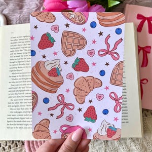 May include: A pink and white patterned card with illustrations of croissants, waffles, strawberries, blueberries, and stars. The card has a pink ribbon and a heart shape.