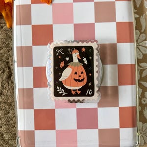 May include: A clear tablet case featuring a pink and tan checkered pattern with a central, rectangular phone grip. The grip displays a black postage stamp illustration of a white goose wearing a pumpkin costume, accented by small leaves and the number 10.