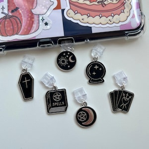 May include: A selection of black and silver charms, including a coffin, a book with "SPELLS", a crescent moon, a crystal ball, a sun and moon, and tarot cards. They are attached to clear plastic clips.