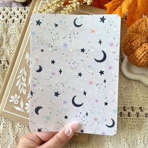 May include: A rectangular card with a celestial design featuring black crescent moons, stars, and iridescent glitter on a white background. The card is held in a hand, with a neutral background of autumn decor.