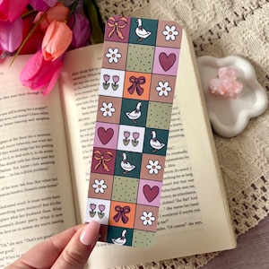 May include: A patterned bookmark with a patchwork design featuring bows, flowers, hearts, and geese. The bookmark is held over an open book, with pink and purple flowers in the background. The design includes squares of various colours.